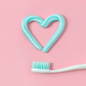 A heart shape drawn with toothpaste sitting above a toothbrush against a pink background